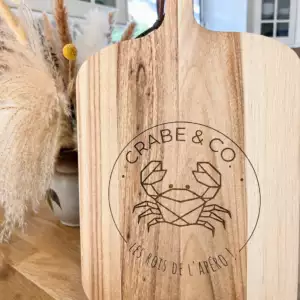Planche ap&eacute;ritive en bois grav&eacute;e au laser avec logo crabe personnalis&eacute; &ldquo;Crabe & Co&rdquo;, pos&eacute;e sur une table en bois dans une ambiance chaleureuse et naturelle.